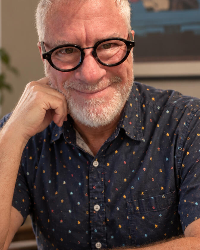 Arden McKenna, contemporary MM romance author Arden McKenna smiling at a writing desk, wearing round black glasses and a navy patterned shirt.